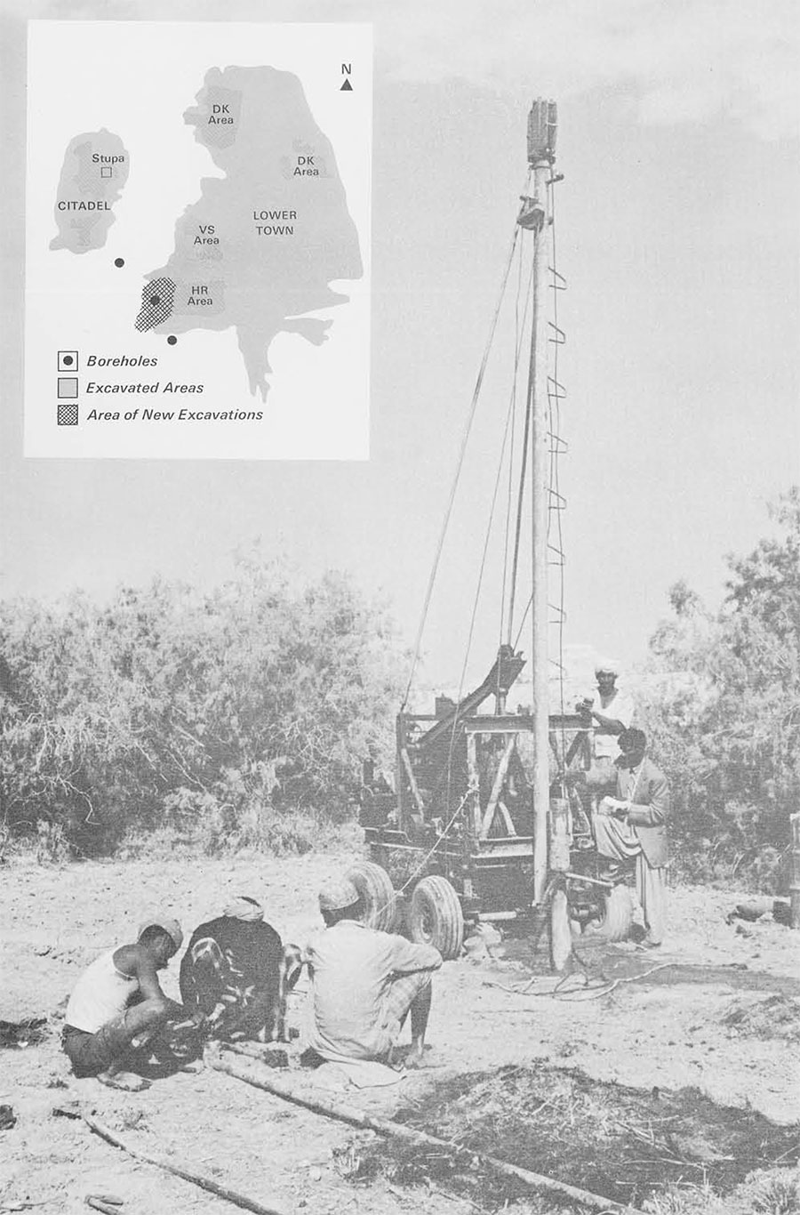 A rig to bore holes in the ground, a group of people stand nearby.