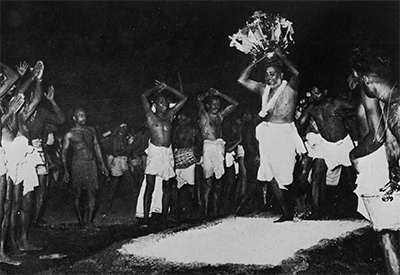 A man holding a vessel above his head as he walks over hot coals, a crowd watches with their arms raised above their heads.