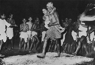 A man holding a child on his hip as he walks over hot coals, a crowd watching.
