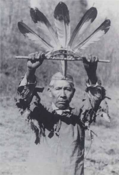 A man holding a feather ward over his head, the wand made of a wooden frame with five feathers fanning above it.
