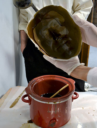 Hands holding a plaster mold with wax coating it, held over a pot of hot wax.