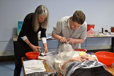 Two people making a plaster cast of the face of a person on a table.