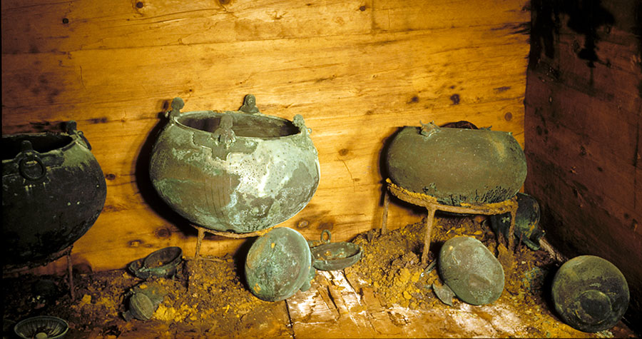 Three large bronze cauldrons on stands, against a wood wall.