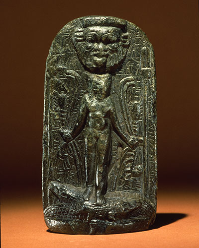 Stela with Child Horus standing on 2 crocodiles.He holds serpents and scorpions in his hands. Head of Bes is above Horus. Inscribed on the back with magic spell.