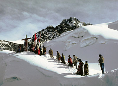 A large group of people trecking up a snowy mountain side, some bearing a large cross and flags.