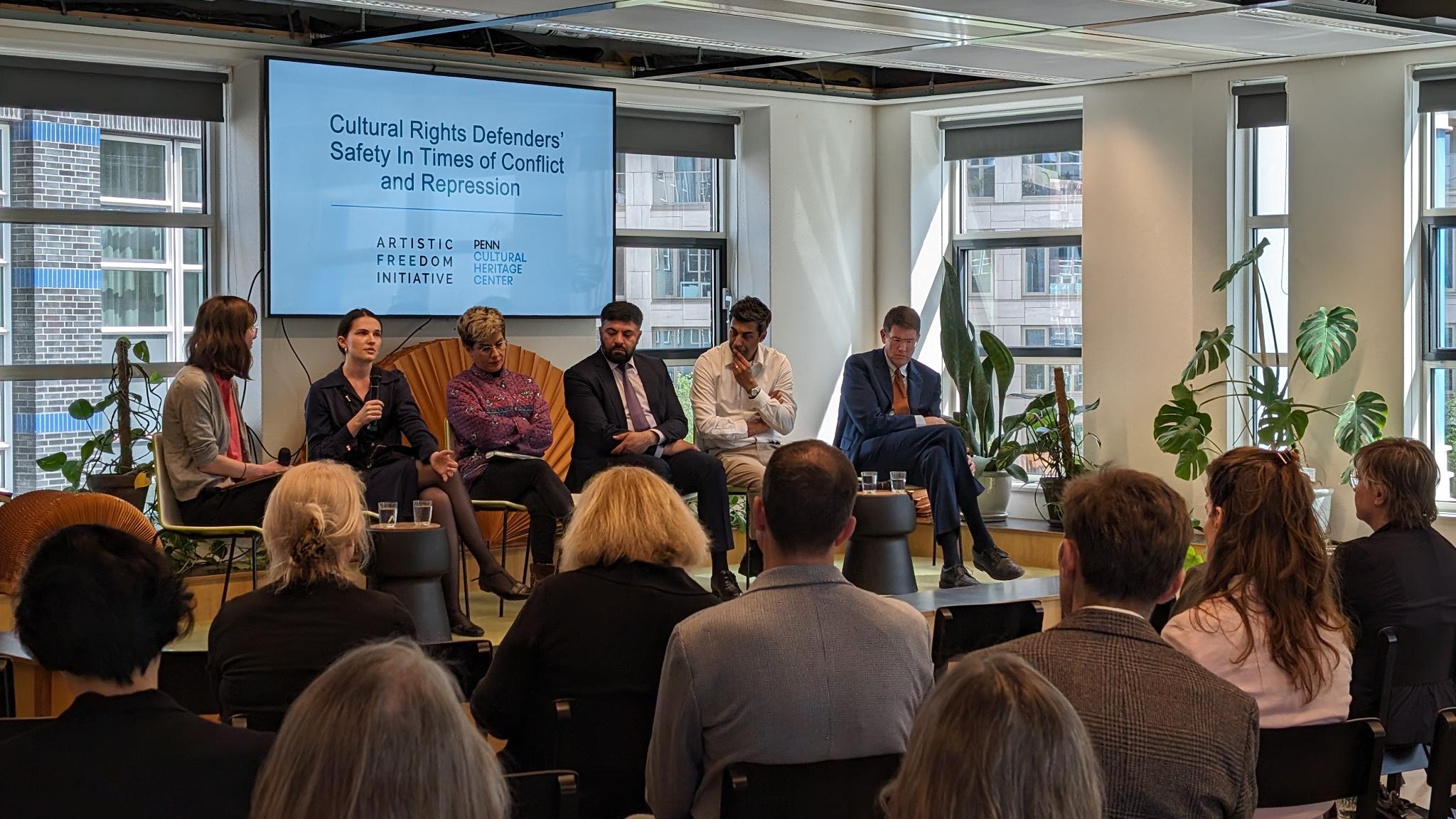 Panel discussion on "Cultural Rights Defenders' Safety in Times of Conflict and Repression" taking place in a conference room with six speakers seated in front of a large window, addressing an attentive audience.
