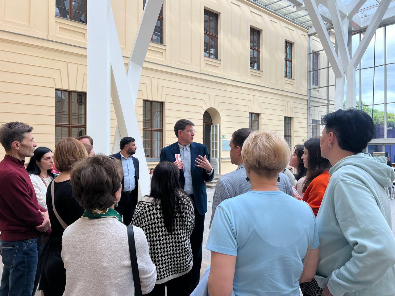 A group of people attentively listens to a speaker, standing in an covered courtyard with a modern, geometric canopy structure and the facade of a classic building in the background.