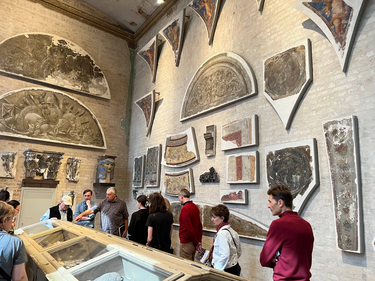 Visitors explore a historic room adorned with ancient fresco fragments at the Neues Museum in Berlin, examining displayed artifacts.
