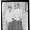 Two women standing, holding hands in front of steps. Both are dressed in late Victorian dress. Haines, Alaska. 1915