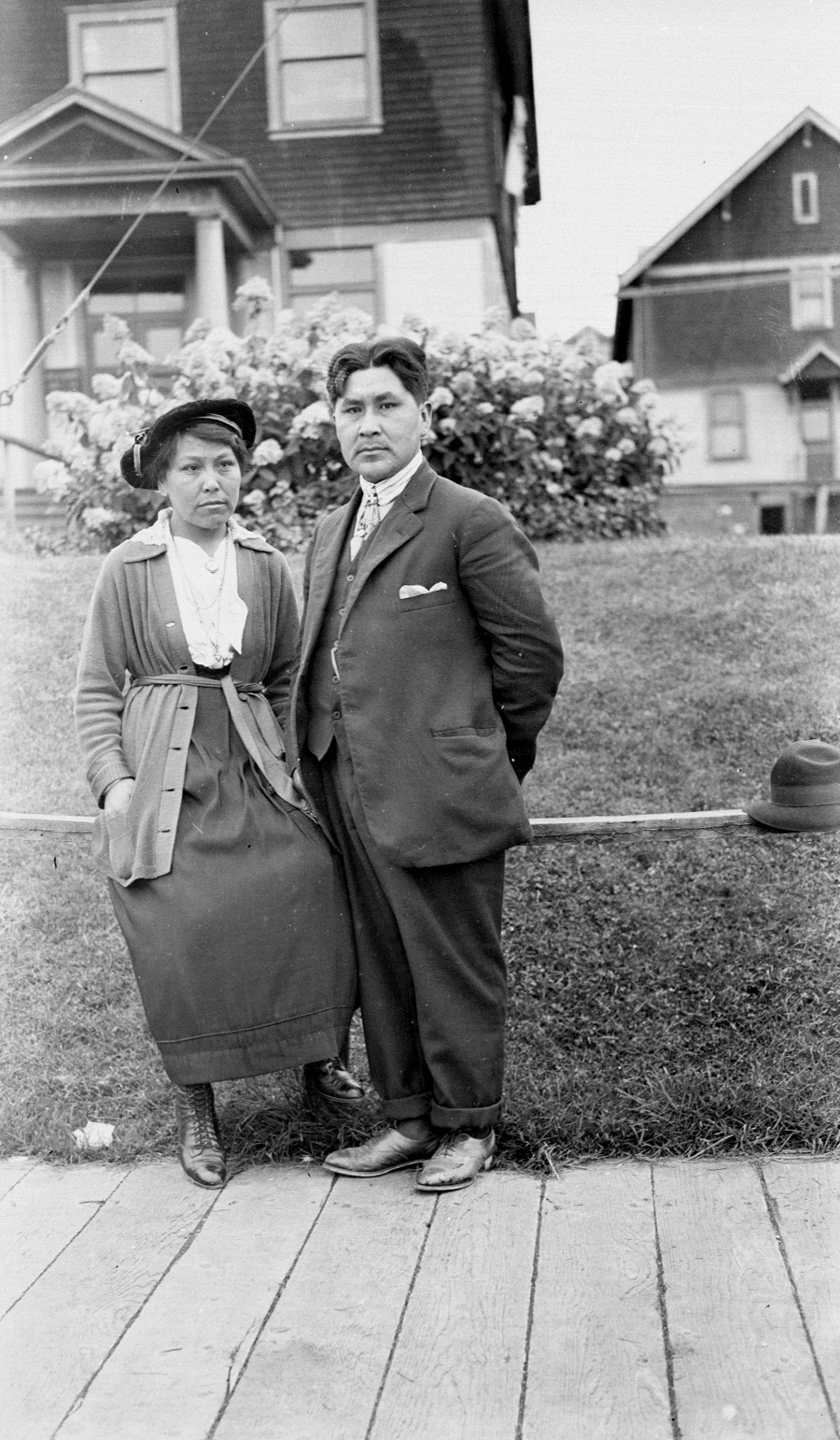 Kwakiutl Man and Wife standing on railing in front of lawn leading to modern houses. Wooden sidewalk. Sept 18, 1918