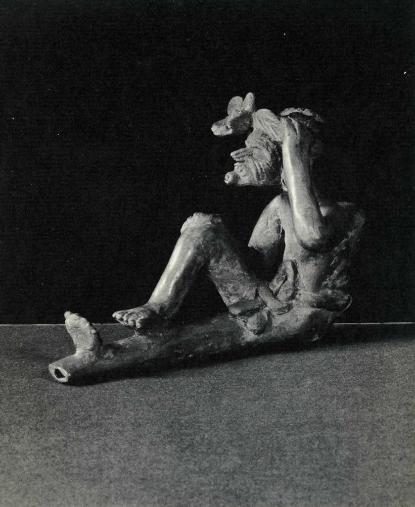 A clay pipe with a seated man wearing a headpiece with a flower decorating the top of the pipe