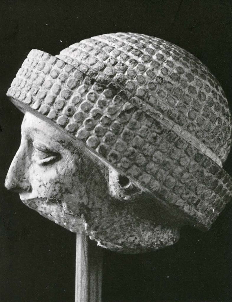 Profile view of a stone carved head, bottom half of the head missing, wearing a textured turban 