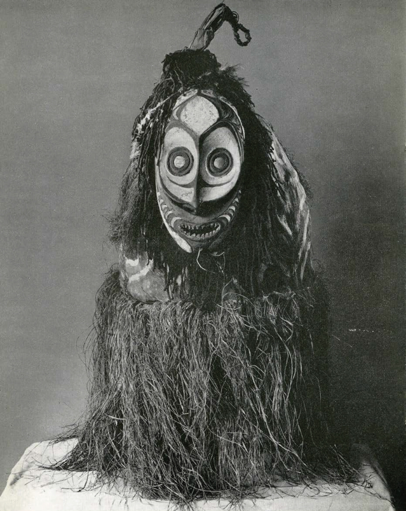 Ceremonial mask that has an open mouth with representation of teeth and is fringed all round with hair, shell inlay eyeballs, around the bottom fringe is of dyed grass