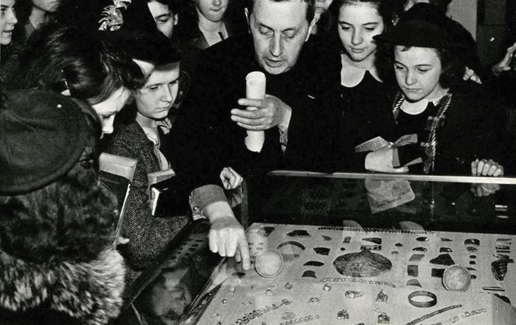 School children grouped around a glass display case full of jewelry, an instructor pointing at an object