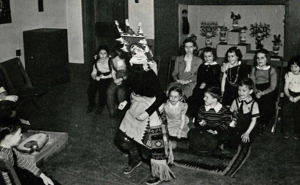 A group of school children watching a dressed up woman perform a dance