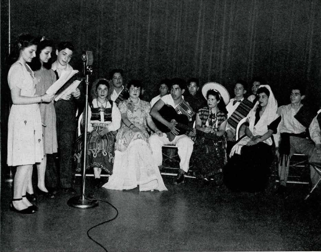 Three people with sheet music singing into a microphone whilst a seated group listens