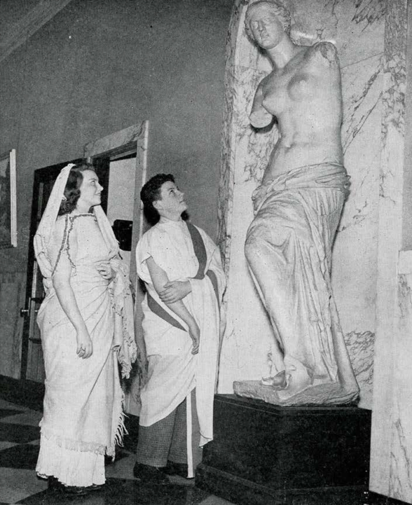 Two women dressed in Roman costumes looking at a sculpture