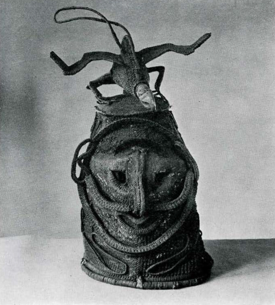 A dance costume headpiece made of basketwork, in front a representation of features of human face; long curved nose; ring marking outline of face, On top a wood and basketry figure of a bird.
