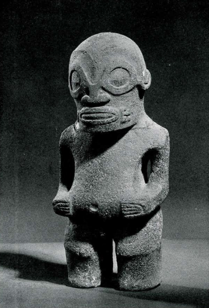 Stone carving of a standing figure with hands on its belly, stylized face.