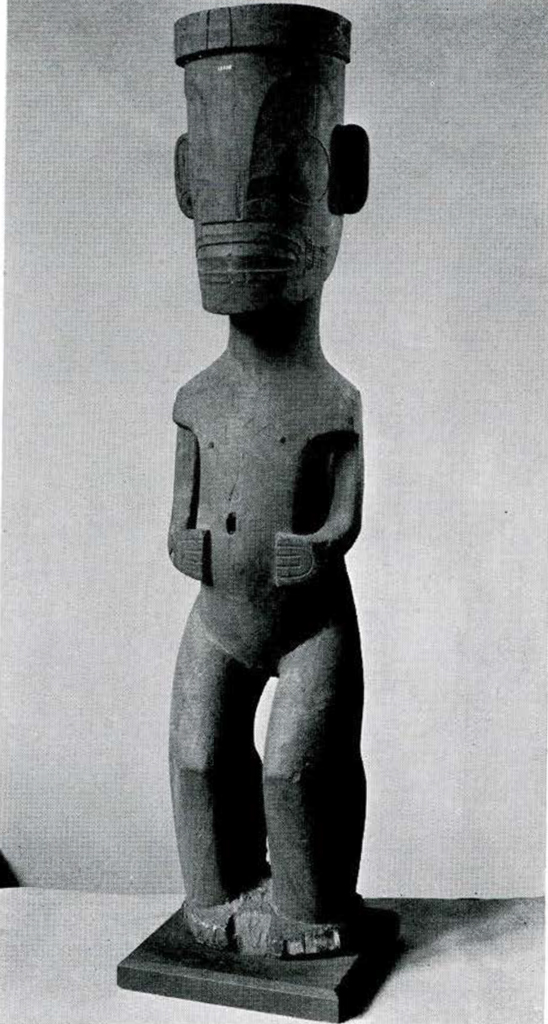 Large wood tiki of a human figure with stylized face, flexed knees, hands resting on stomach.