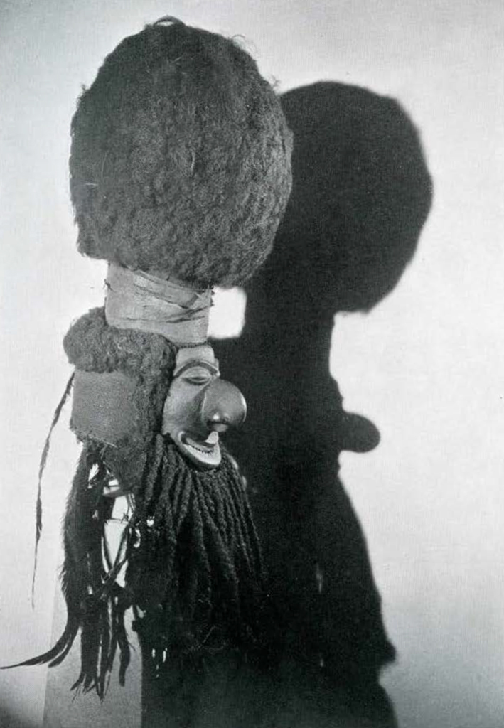 Profile view of a wood mask with large round nose, a large topknot of hair and long beard maid of braided strands