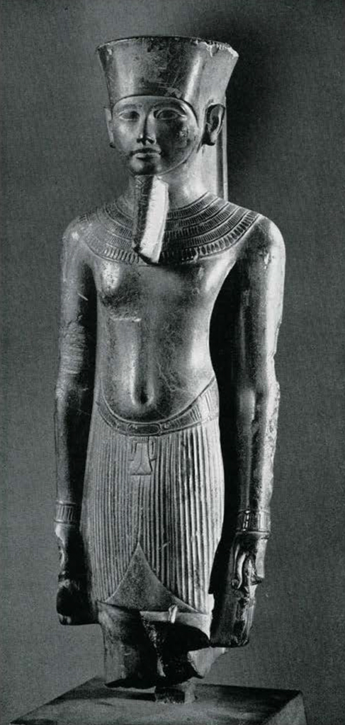 Standing graywacke statue of Amun, preserved from the knees up, wearing a short kilt fastened with a tyet amulet, and a broad collar, the tall plumes of his crown are missing, in his hands he holds two ankh symbols.