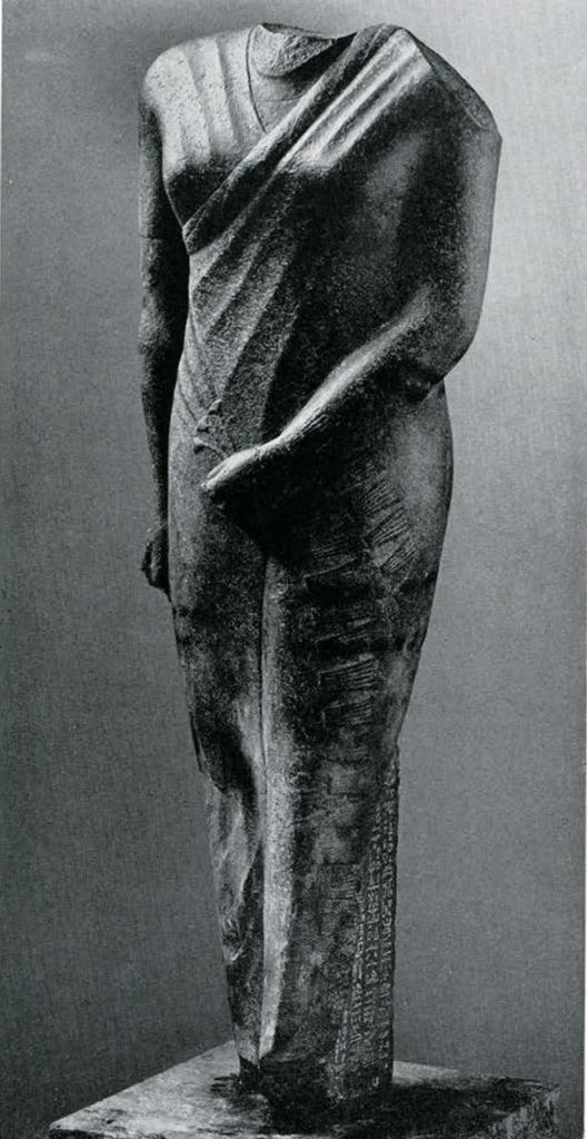 Standing Headless statue of a man wearing a tight fringed garment and holding 3 papyrus blossoms in left hand, head and feet missing. 