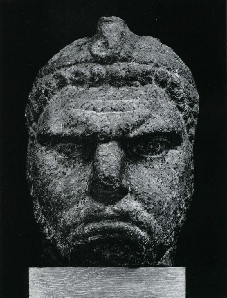 Colossal Head of block-like shape with scowling visage and a rectangular back pillar, hair is treated with bead-like globules arragned in rows framing forehead and continuing on sides and top of head, a wide flat diadem with the uraeus at the front encircles the head, the short beard is treated the same way as the hair.