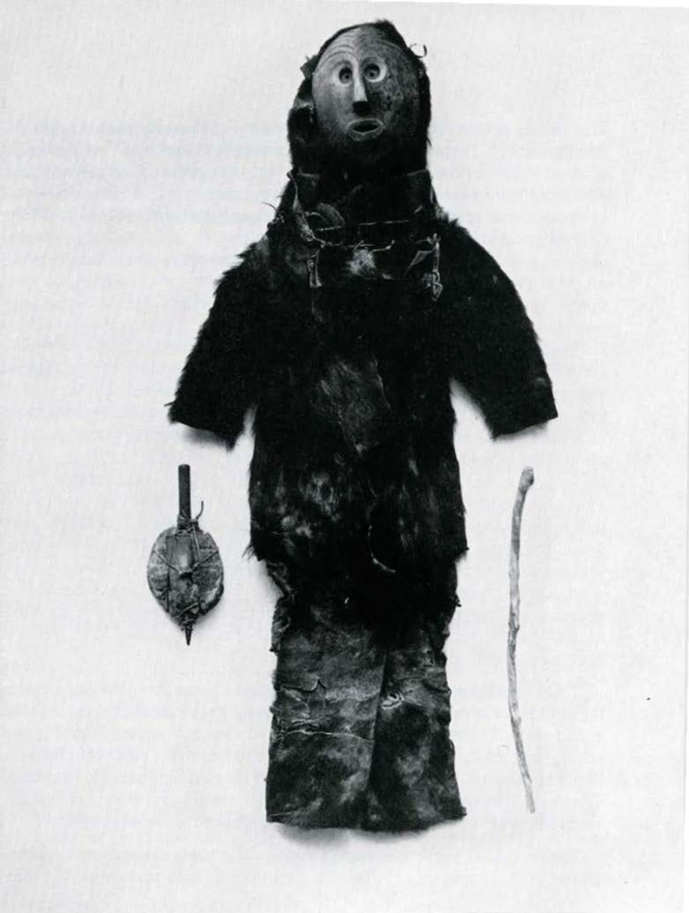 A full body costume made of bear skin, a rattle and cane, and a wood mask with wrinkles and close set eyes.