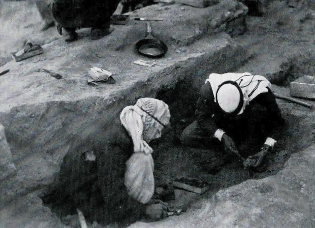 Two men digging in a rectangular pit, removing tablets.