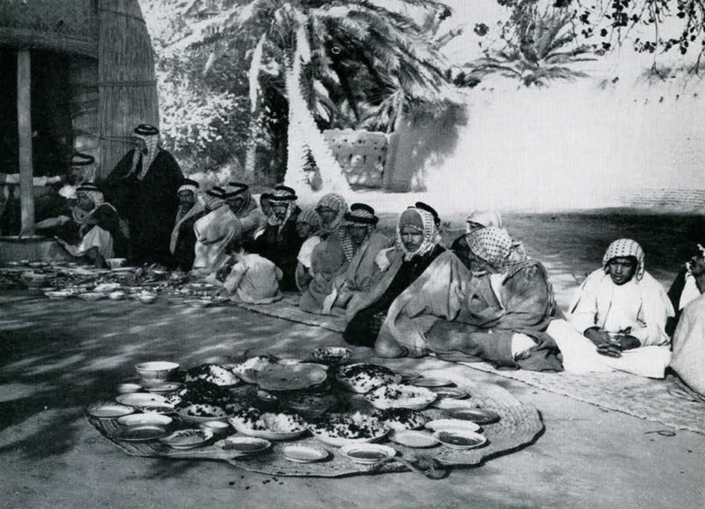 A circular mat table filled with bowls and plates of food, a group of people sitting around it.