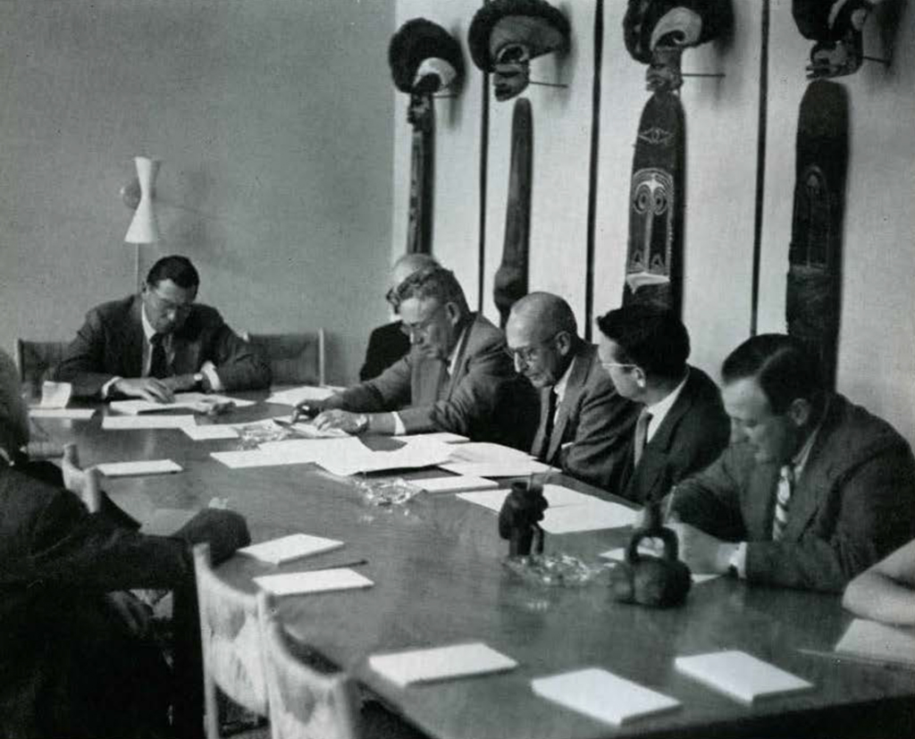 The board of managers at a large table full of papers, artifacts hanging on the wall behind them.