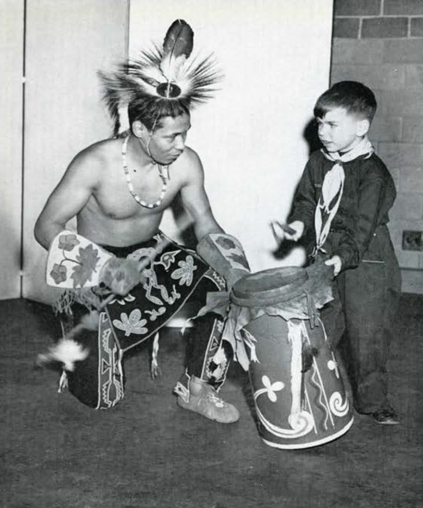 A young boy beating a drum with a kneeling man dressed in his native garb.
