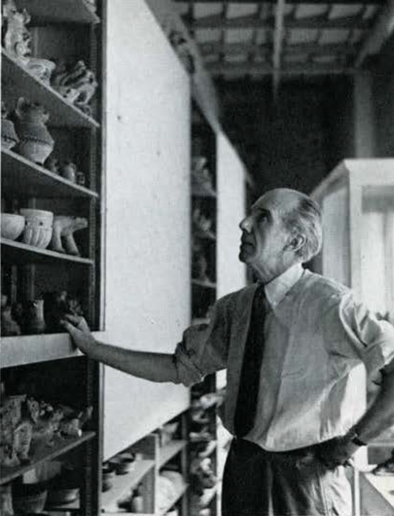 A man looking at a book case full of pottery objects.