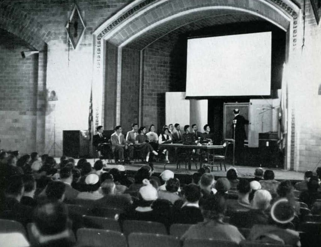 A audience seated in an auditorium, watching a lecturer on the stage.
