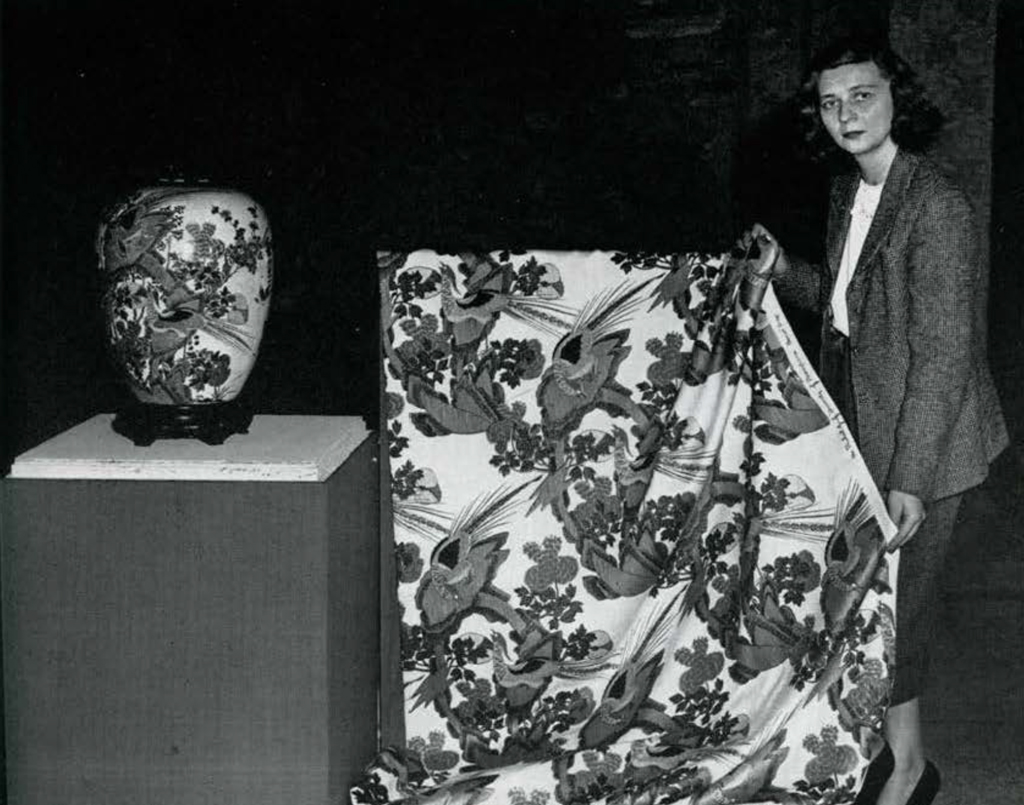A woman holding a bolt of fabric with the same design on it as the vase on a pedestal next to it.