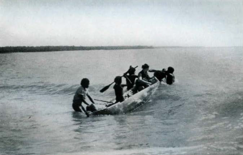 A group of people getting into and paddling a canoe into an oncoming wave.