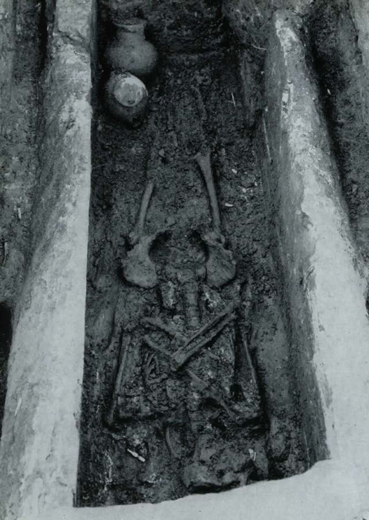 A skeleton with crossed arms in a tomb, two pots at the feet.