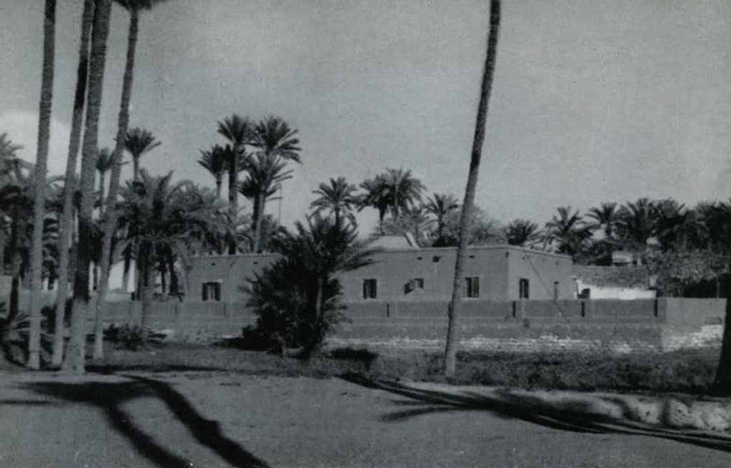 The expedition house, a one level building with a low lying wall around it, palm trees surrounding.