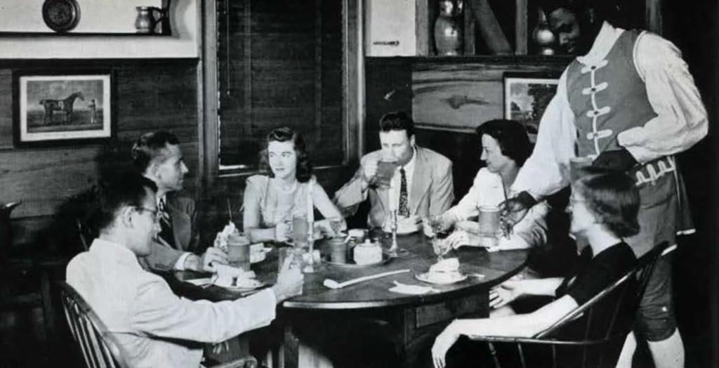 A group of people having a meal at a round table, behing served by a person in period dress.