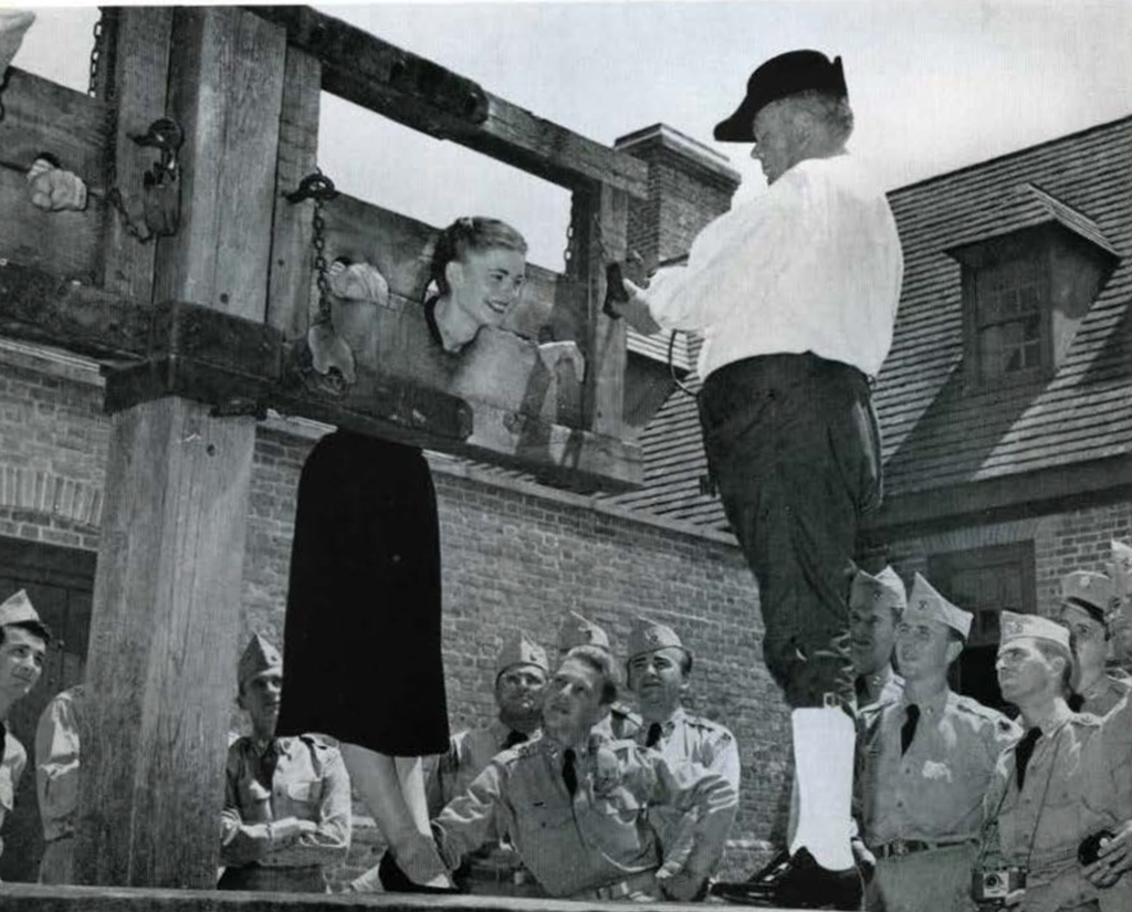 A young woman in mock stocks, a man in period dress in front of her, a crowd of soldiers watching.