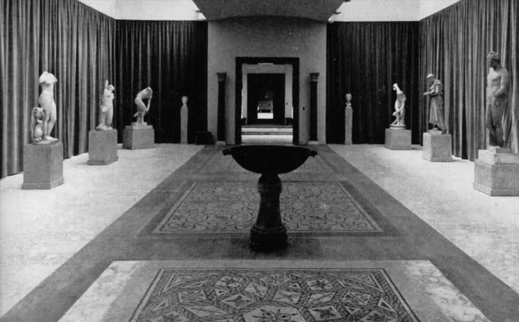 A gallery lined with statues on pedestals, the floor has large mosaics.