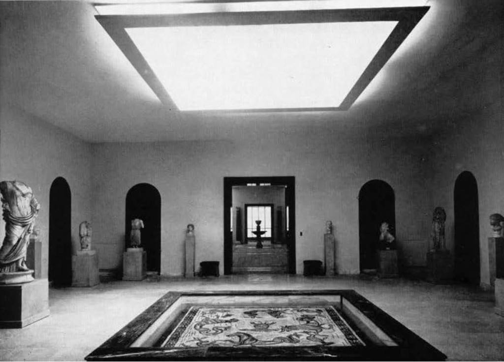 A gallery with statues on pedestals lining the walls, a large mosaic in the middle of the floor.