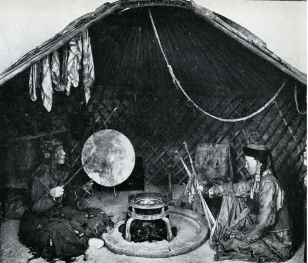 Two people, seated on the ground with a firepit between them, one person banging on a drum.