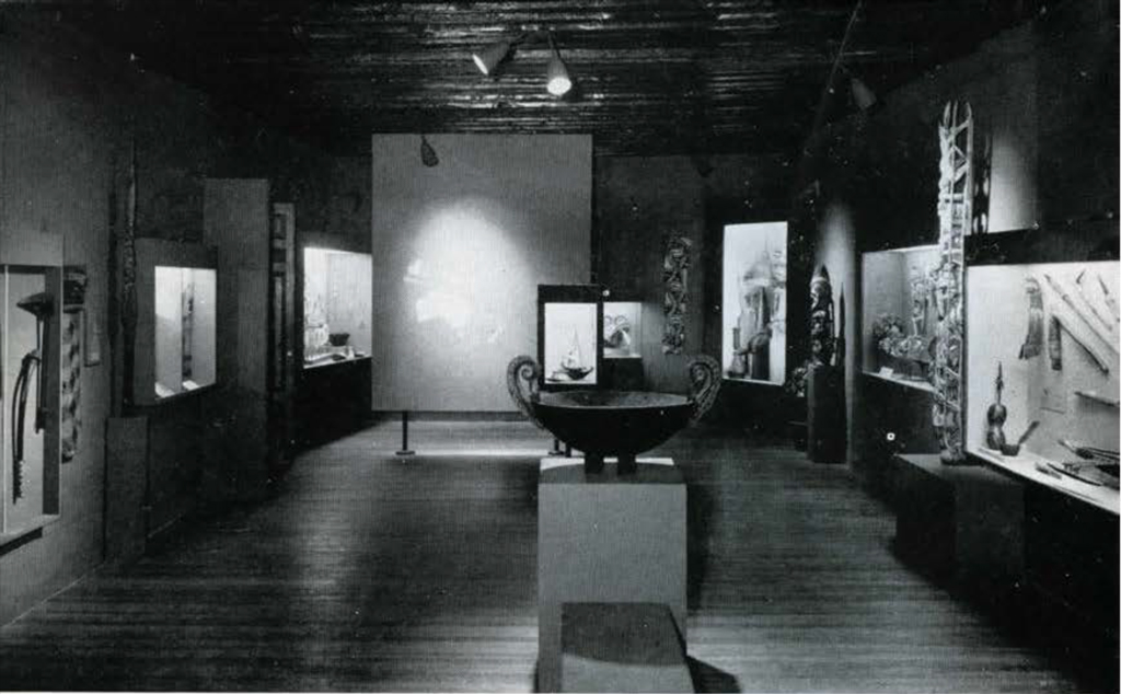 A museum gallery with display cases showing objects from the Pacific, a vase on a pedestal in the middle of the gallery.