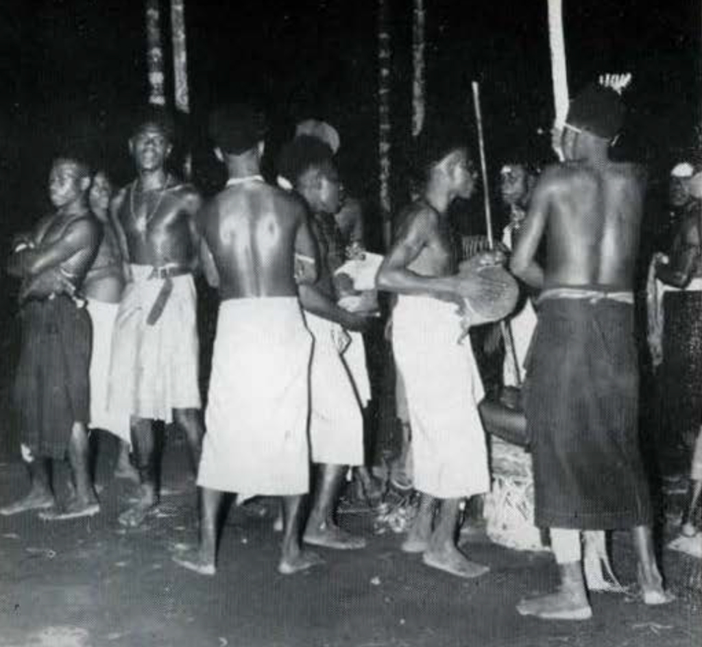 A group of men wearing skirts, some hold spears.