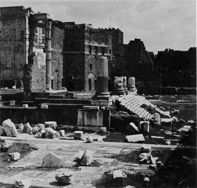 The forum of Augustus, large pieces of rubble strewn about, stairs and intact buildings in the background.
