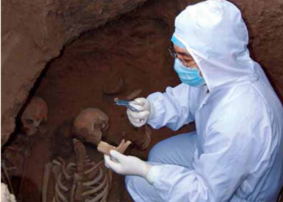 A scientist in a white jumpsuit and mask collecting bone samples from a burial site, remains and vessels in the burial.