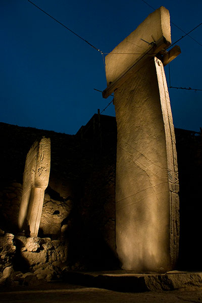Massive stone pillars lit up at night, showing collosal carvings on arms.