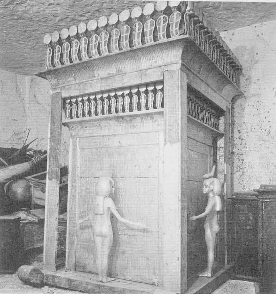 A tall and ornately decorated box with a figure standing on each side, facing towards the box, arms open to emcompass the box.